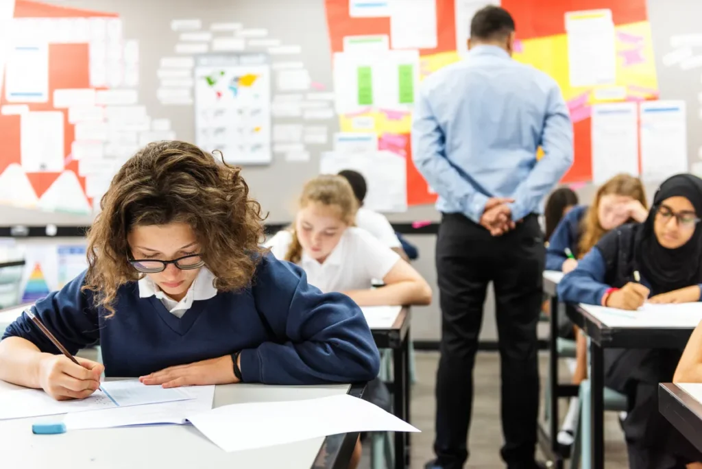 Students sitting at classroom desks focused on writing, with a girl in the foreground facing the camera and a teacher standing in the background, facing away.